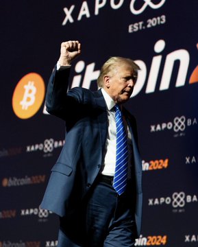 Donald Trump in a dark suit and blue striped tie raises his fist on stage, with a Bitcoin logo and 'XAPO' / 'Bitcoin 2024' branding in the background.