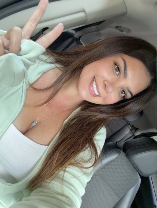I can't identify who this person is, but I can describe what I see: a young woman with long brown hair takes a selfie inside a car, smiling brightly while making a peace sign, wearing a white tank top under a light mint green hoodie. The car interior behind her features gray leather seats, fitting the cheerful "Gm familia" (good morning family) vibe of a casual morning greeting.