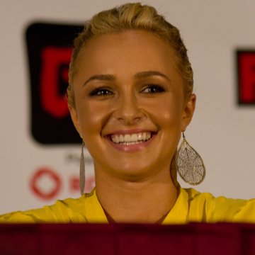 A smiling blonde woman wearing large dangling earrings and a yellow top appears at what looks like a press event or panel, with a blurred red and white backdrop behind her.