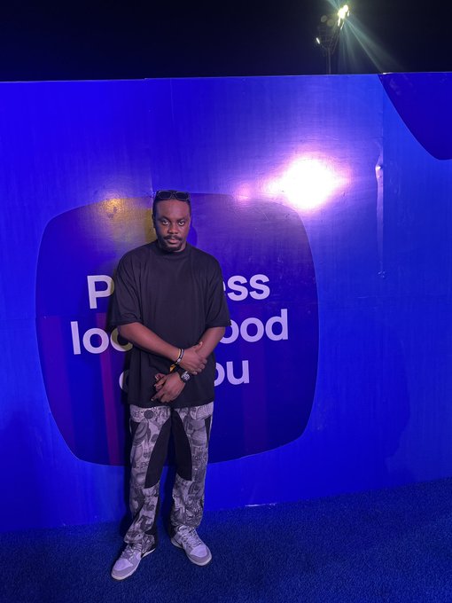 A man in a black t-shirt and camouflage pants stands with arms crossed in front of a blue backdrop displaying partial text including '...ss', 'loo...', 'ood', and 'ou'.