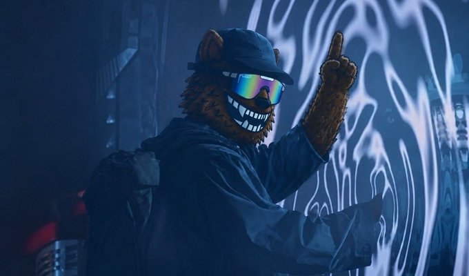 An anthropomorphic bear in a dark hoodie, cap, and reflective sunglasses points upward while standing in front of a graffiti-covered wall.