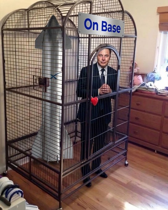 A man in a suit stands inside a large wire cage labeled 'On Base' next to a rocket model and an inflatable shark, with a red heart attached to the bars.