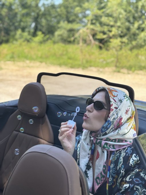 A woman in a headscarf blows bubbles while riding in a convertible, reflecting a sense of carefree freedom. The text "DARS" is visible on her scarf.