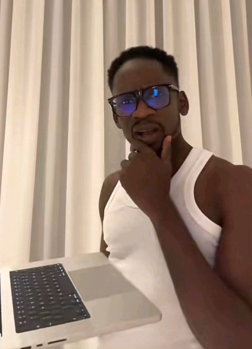 A thoughtful-looking man wearing blue-tinted glasses and a white tank top sits in front of white vertical blinds with a laptop in front of him, resting his hand on his chin in a contemplative pose that mirrors the pondering tone of the post about the psychology behind why Web3 creators reveal their identities on TikTok but remain anonymous on X.