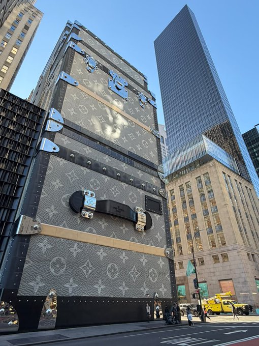 A massive building facade designed to look like a giant Louis Vuitton trunk with monogram patterns and silver hardware, situated in an urban setting with other skyscrapers.