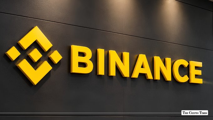 A photograph of the yellow Binance logo and 'BINANCE' text mounted on a dark wall, with 'THE CRYPTO TIMES' watermark in the corner.
