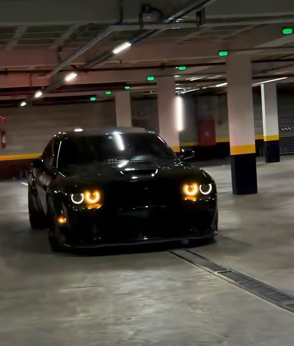 A black Dodge Challenger with its signature halo headlights illuminated is parked in an indoor parking garage.