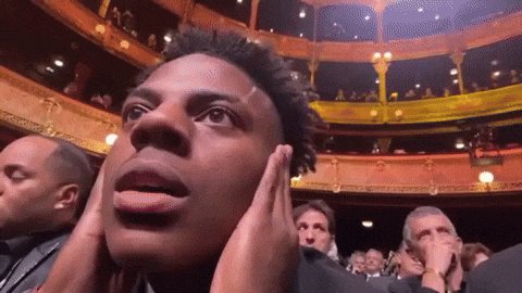 The image shows a man with dreadlocks covering his ears while looking up at something or someone above him. The scene takes place inside an auditorium filled with people who appear to be watching a performance, possibly a concert or a play. The audience is seated in rows of chairs and appears to be engaged in the event. The man's reaction suggests that he might be reacting to a surprising or unexpected occurrence during the performance.