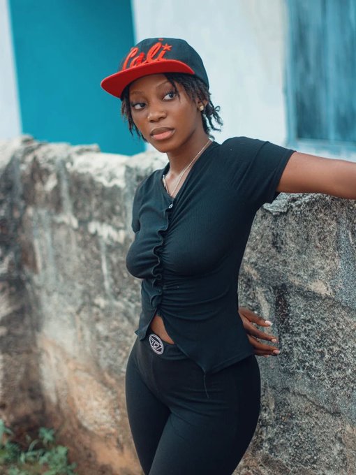 A portrait photograph of a young woman wearing a black "Cali" baseball cap, black ruffled top, and black pants, posing against a stone wall with a teal-colored background.