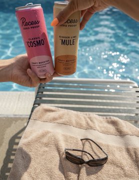 Two hands clink cans of Recess "Cosmo" and Recess "Ginger Lime Mule" zero-proof sparkling waters over a poolside lounger, with sunglasses and a towel resting below. The leisurely scene embodies the post's mantra—holding quality investments ($TROLL) to enjoy stress-free, indulgent moments like this.
