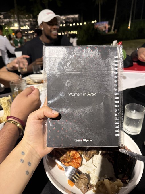 A person holding a dark, patterned notebook titled 'Women in Avax' and 'team Nigeria' over a plate, with people visible in the background at an outdoor nighttime event.