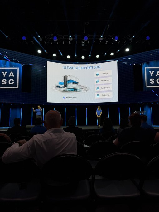 An audience watches a presentation in a large conference room, where a screen displays 'ELEVATE YOUR PORTFOLIO' with the Yardin Wealth Group logo and a list including Leasing, Operations, Construction, and Budgeting.