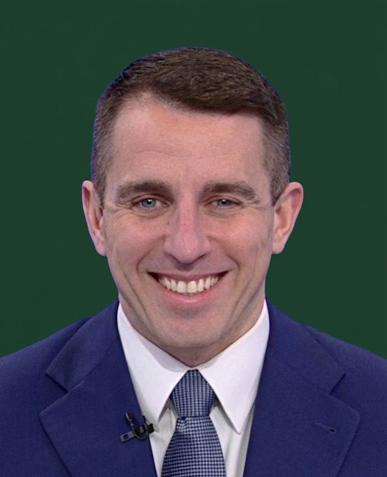 Professional headshot of a smiling man in a navy blue suit, white shirt, and patterned blue tie against a dark green background, wearing a small lapel microphone.