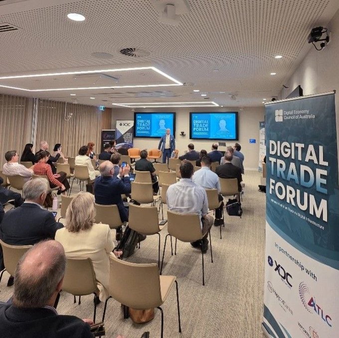 A conference room filled with attendees watching a presentation at the Digital Trade Forum, organized by the Digital Economy Council of Australia with banners and screens displaying the event name.