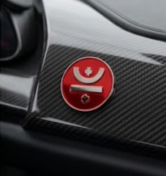 A close-up of a red circular emblem with a stylized symbol mounted on a carbon fiber surface, likely part of a vehicle.