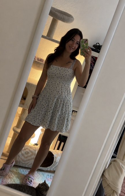 A mirror selfie of a young woman with long dark hair wearing a white floral sundress and pink Crocs, taking a photo in her bedroom.