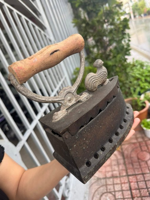 An old-fashioned metal charcoal iron with a wooden handle and ventilation holes, held outdoors against a background of plants and a tiled floor.