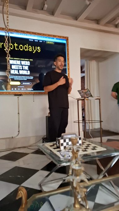 A man in black clothing stands speaking in front of a screen displaying 'crypto.days WHERE WEBS MEETS THE REAL WORLD,' with a chessboard on a table nearby.