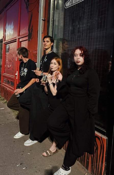 Four people dressed predominantly in black stand and lean against a red building exterior at night, with one person smoking a cigarette.