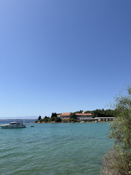 A coastal landscape with clear turquoise water, a small white boat, and buildings on a peninsula under a bright blue sky.