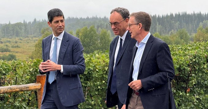 Three men in business suits standing outdoors in front of a forested area, appearing to be in conversation during what looks like a formal outdoor meeting.