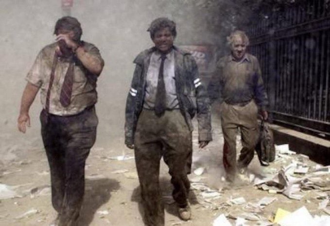Three men in business shirts and ties, covered in dust and debris, walk away from the 1993 World Trade Center bombing site through a smoke-filled street littered with paper.