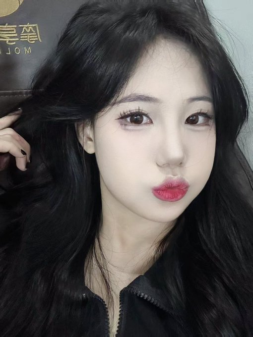 A close-up selfie of a young woman with long dark hair, wearing pink eye makeup and bright pink lipstick, posing with puckered lips and one hand near her hair.