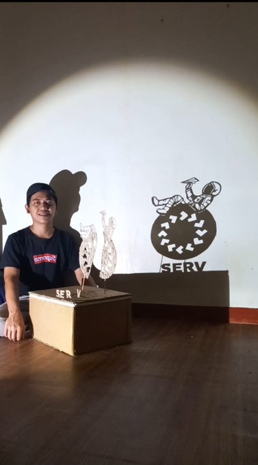 A smiling man in a black t-shirt and cap sits next to a wooden pedestal displaying a detailed wire sculpture, with a wall logo reading 'SERV' behind him.