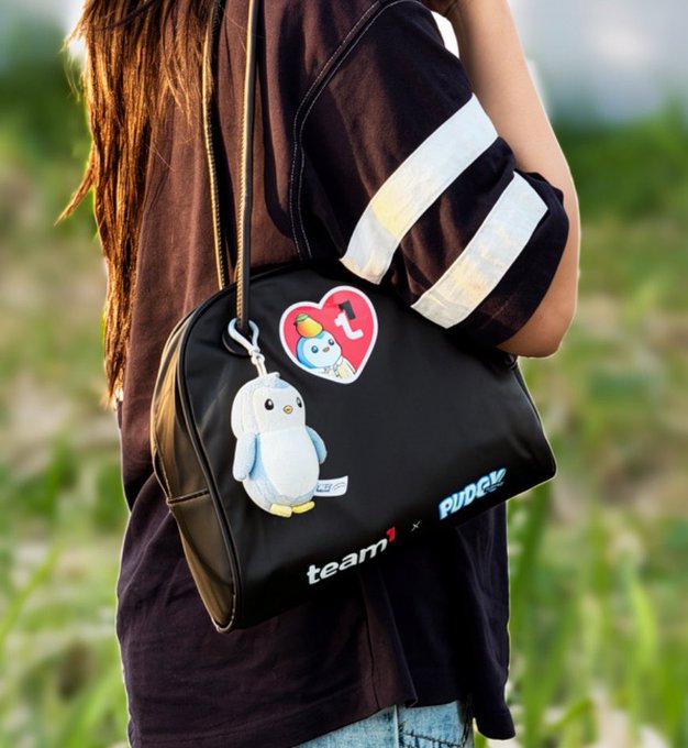 A person carries a black bag with 'team1 x PUDGY' branding, featuring a Pudgy Penguin plush keychain and a heart sticker.