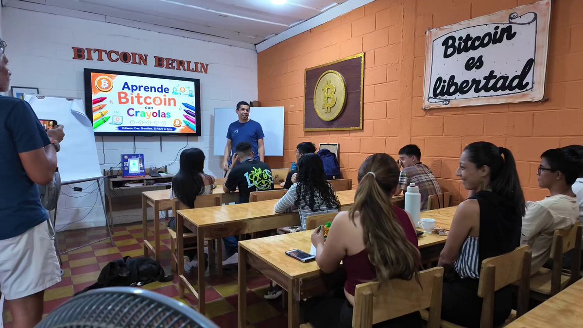 The image shows a group of people sitting around tables in front of a television screen displaying information about Bitcoin. A man stands behind them, likely giving a presentation or lecture on the subject. The room has brick walls and wooden floors, creating an educational atmosphere for learning about Bitcoin.