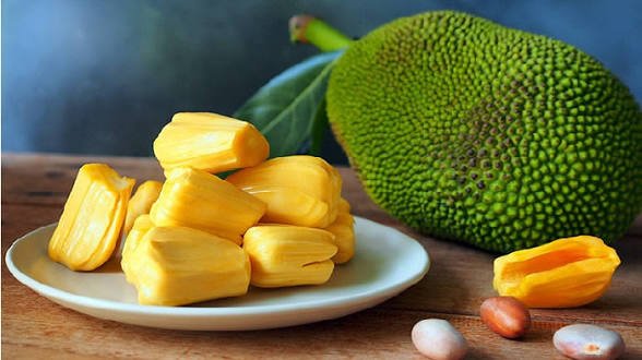 A white plate holds fresh, bright yellow jackfruit bulbs alongside a whole green jackfruit on a wooden surface, with a few seeds visible in the foreground. The image perfectly illustrates the post's "Jackfruit party" theme, showcasing the tropical fruit being enjoyed in the office as a morning treat.