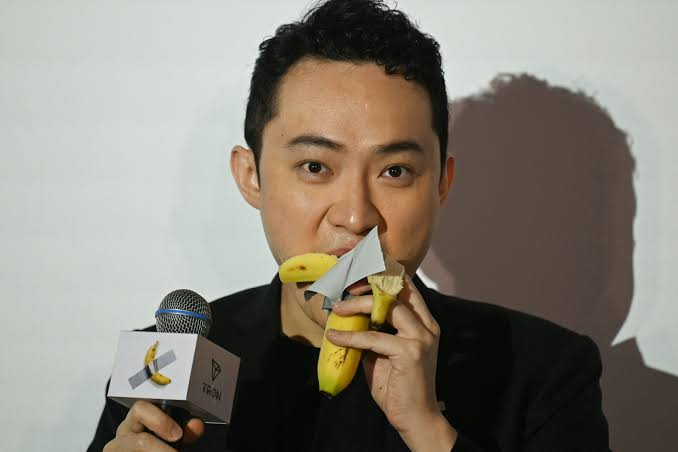 A man wearing a black jacket holds a microphone with a TRON logo and a small object wrapped in gray tape with a banana emoji, biting into a banana.