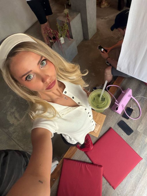 A selfie of a young blonde woman taking a morning photo at a café, with a green matcha drink, a pink purse, and a phone on the table beside her, captioned "Good morning legends. Lets have a great day." to match the cheerful, start-of-day vibe.