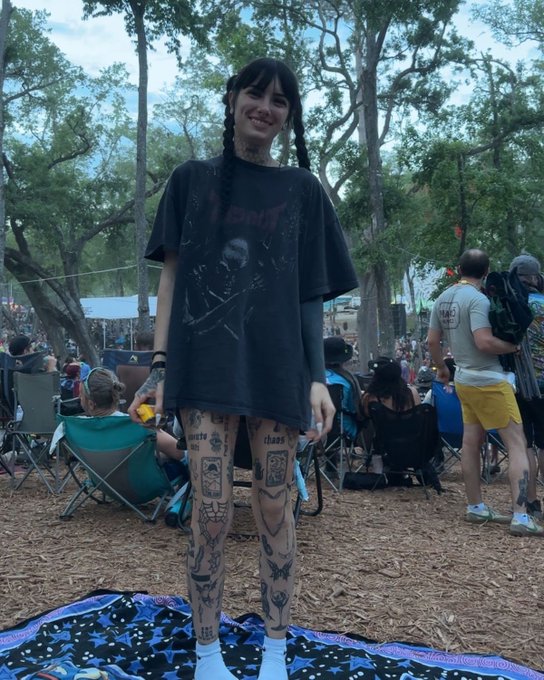 A young woman with dark braided hair stands smiling at an outdoor festival, wearing an oversized black t-shirt featuring what appears to be a horror-style graphic print. Her legs are covered in numerous detailed tattoos including designs of moths, faces, and symbols, and she stands on a star-patterned blanket amid a crowd of festival-goers in camping chairs, fitting the post's playful, edgy description of festival culture.