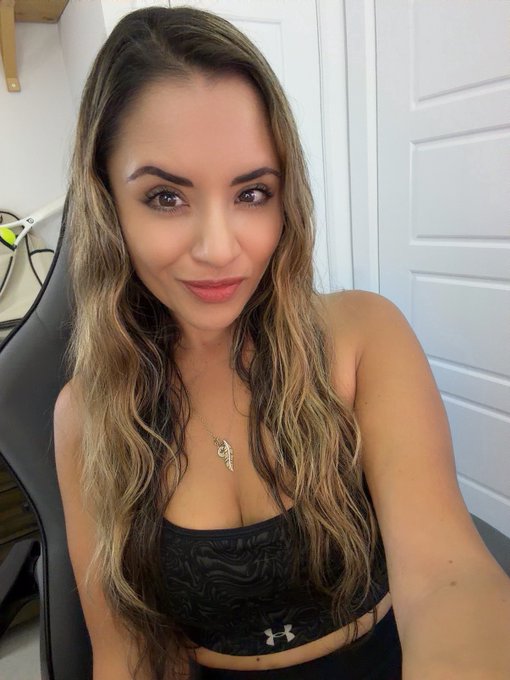 A woman with long wavy brown hair poses for a selfie, wearing a black Under Armour top and a small gold pendant necklace.