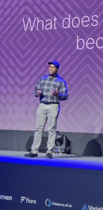 A man wearing a blue cap, plaid shirt, and khaki pants speaks on stage at XRP Seoul 2026, with the partially visible presentation text "What does beco..." displayed behind him on a purple patterned backdrop, consistent with the event's branding shown at the bottom including logos for Flare, DataVault.ai, and Varia.