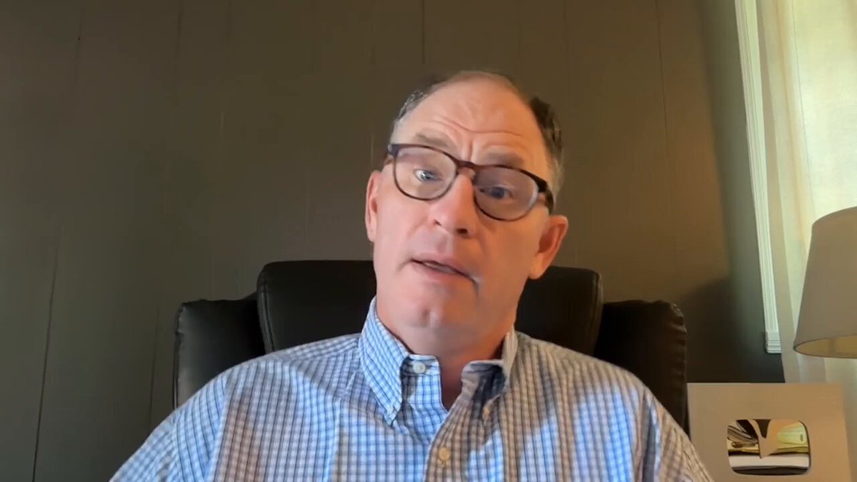 The image features a man wearing glasses, sitting in an office chair with his hands together at his sides. He appears to be engaged in a video call or participating in a live stream event. The background of the image shows a wall that has a window on it, suggesting that he might be indoors and possibly working from home. There is also a lamp visible in the scene, which could indicate that the man is using natural light during his workday.