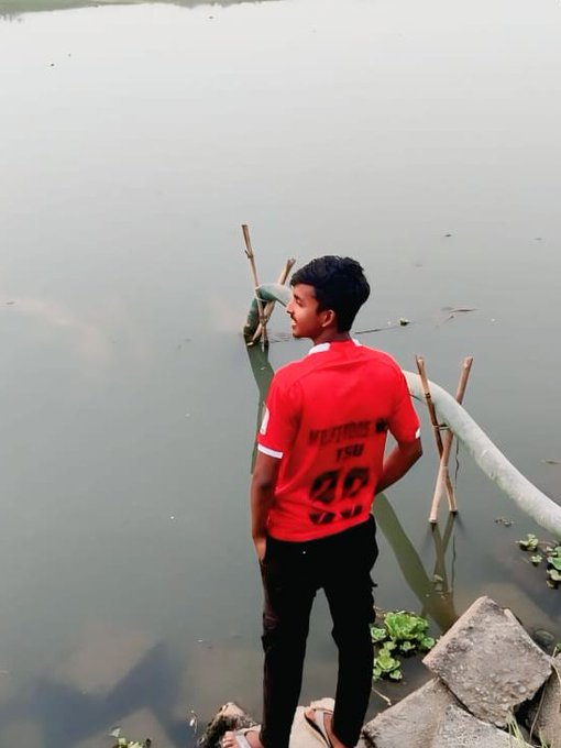 The image shows a young man standing near some pipes that are floating in water, possibly at a lake or river. The man appears to be wearing a red shirt with the number "48" printed on it. He seems to be looking out over the water, possibly contemplating something or enjoying the view.