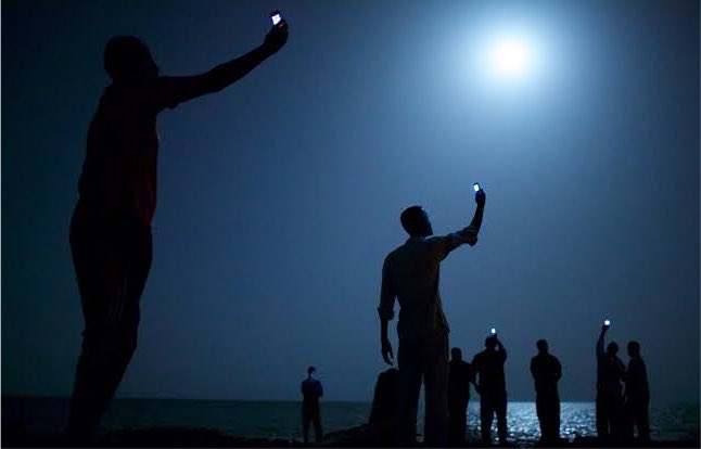 A nighttime silhouette photograph showing several people on a shoreline holding up small lights, with a bright full moon glowing in the dark blue sky.