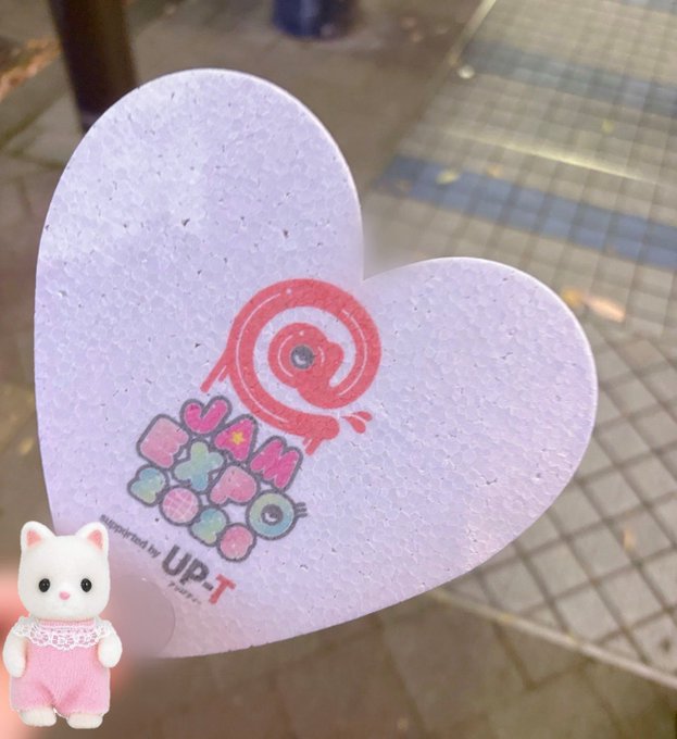 A heart-shaped cardboard sign with a Tamagotchi logo and the text 'Tamagotchi UP.T' lies on a tiled ground, with a small Sylvanian Families cat figurine placed on it.