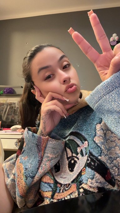 The image features a young woman with long hair, wearing a sweater with a cartoon character design. She is posing for a selfie by making a peace sign with her fingers while sitting at a table or desk. The caption of the photo reads "Gmfers... Day 143 of a selfie a day until MON hits ATH Btw... In order for good things to come your way you must believe that you deserve them". This message seems to be related to the post about the importance of self-love and positivity, as suggested by the caption.