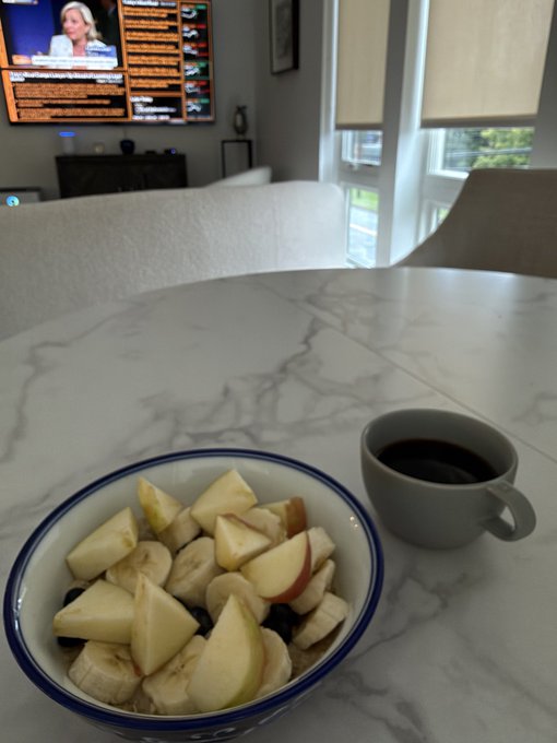 A bowl of sliced apples and bananas with a cup of dark liquid sits on a marble table in a living room, with a television displaying a news broadcast visible in the background.