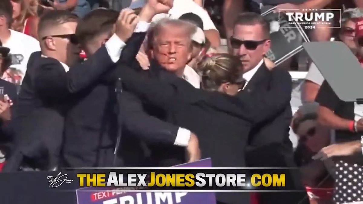 A news screenshot from 'THEALEXJONESSTORE.COM' showing Donald Trump being escorted by Secret Service agents with raised fists at a campaign rally, with a 'TRUMP 2024' logo in the corner.