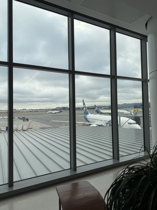 View from an airport terminal window showing Alaska Airlines planes on the tarmac under a cloudy sky, with a yellow "Y4" sign visible near the gate, suggesting travel anticipation that pairs well with the question "Where to?"