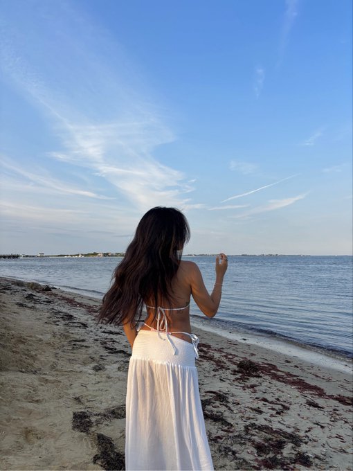 The image shows a woman standing on a beach, wearing a white bikini top and skirt. She appears to be enjoying her time at the beach, possibly taking a break from checking social media or looking at her phone. The photo captures a moment of relaxation for the woman as she looks out towards the ocean.