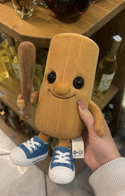 A person's hand holding a plush toy that resembles a wooden baseball bat with a smiling face, blue sneakers, and holding a small brown bat.