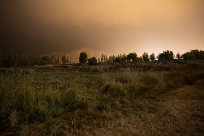 A dusky landscape with a mountain in the background, silhouetted trees, and a grassy field in the foreground under a hazy orange sky.