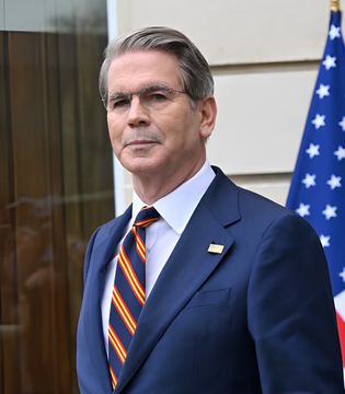 Scott Bessent, U.S. Treasury Secretary, stands before an American flag in a navy suit with a striped tie, pictured as the article discusses how the Treasury could deploy its $1 trillion Treasury General Account via bond buybacks to add liquidity — potentially a catalyst for Bitcoin and broader markets.