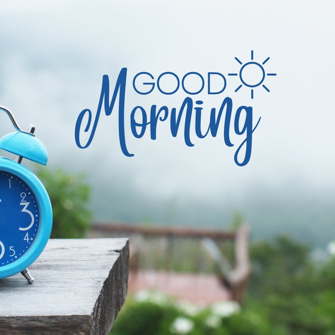 A 'GOOD Morning' greeting graphic featuring a blue alarm clock on a wooden surface with a misty mountain landscape in the background.