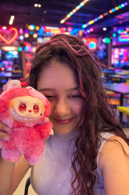 A smiling woman with curly reddish-brown hair holds a pink plush toy in what appears to be a colorful arcade or entertainment venue.